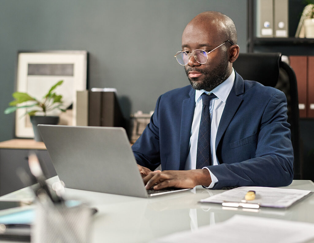 Hombre con gafas, vestido con traje azul, trabaja en una laptop en un entorno de oficina. Se observan plantas y documentos en una mesa clara. Fondo con estanterías y cuadros.