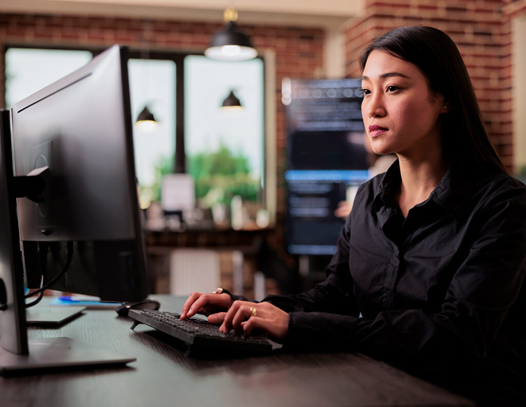 Mujer sentada frente a una computadora, tecleando, en un entorno de oficina moderno con paredes de ladrillo y ventanas grandes que permiten la entrada de luz natural.
