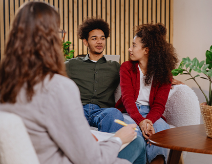 Pareja sentada en un sofá conversando, mientras una tercera persona toma notas en un cuaderno. El entorno es acogedor, con plantas y una pared de madera clara.