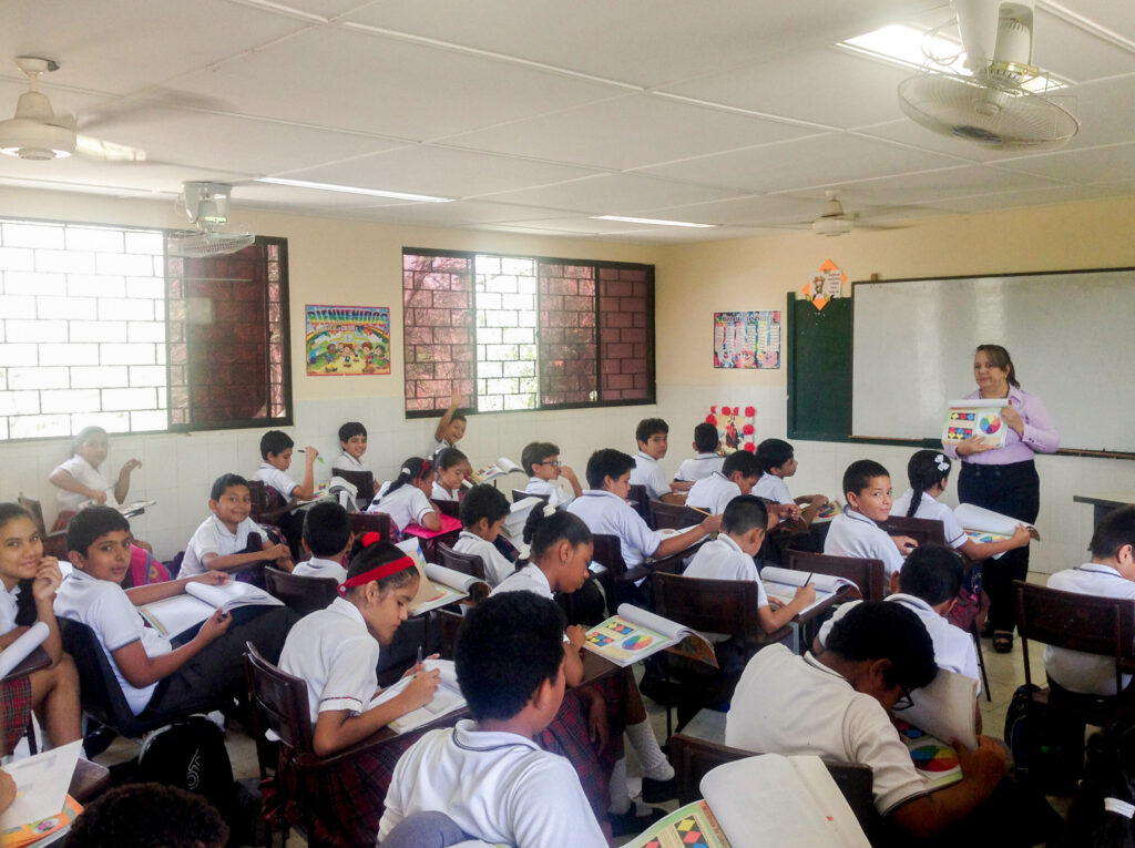 Aula con estudiantes sentados en escritorios, una maestra en el frente sostiene un material didáctico, ventanas con luz natural, paredes decoradas y alumnos escribiendo en sus cuadernos.
