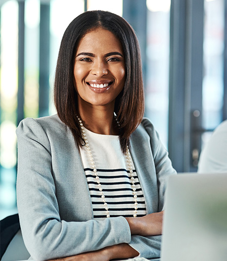 Mujer sonriente con cabello largo y lacio, vestida con blazer gris y blusa a rayas, sentada frente a una computadora portátil en un entorno de oficina con luz natural.
