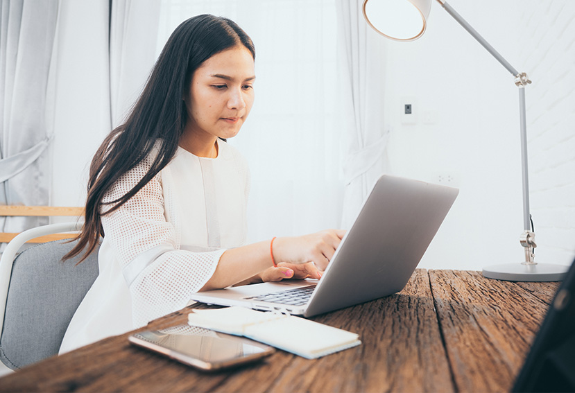 Mujer joven trabajando en una computadora portátil sobre una mesa de madera, con un teléfono móvil y un cuaderno.