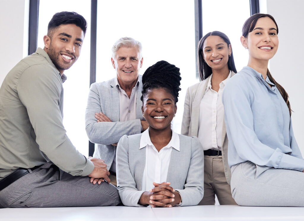 Grupo de cinco personas en un entorno de oficina moderna, posando con sonrisas. Predominan colores neutros en la vestimenta y un fondo luminoso con ventanas.