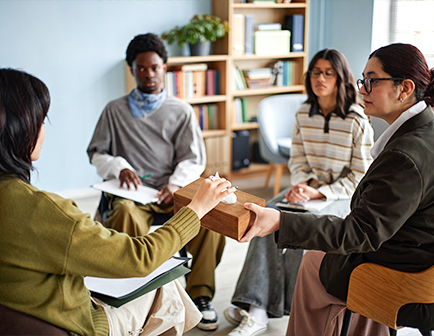 Grupo de cuatro personas en una reunión, una mujer pasa un objeto de madera a otra, mientras los demás observan en un entorno con estanterías y decoración sencilla.